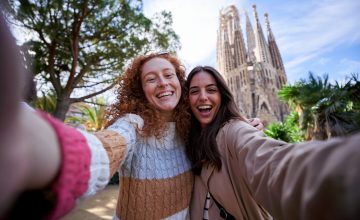 Two happy women friends taking a selfie in front of the sagrada familia, enjoying their vacation together, looking at camera smiling.